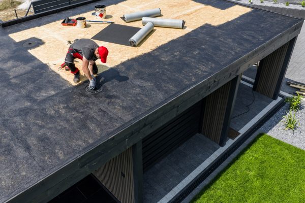 A construction worker is installing EPDM roofing material on a contemporary home under clear skies, surrounded by tools and prepared materials for the roofing project.
